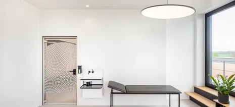 Modernes Behandlungszimmer mit großen Fenstern und einer medizinischen Liege auf grauem Betonboden